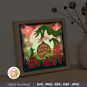 May include: A layered paper-cut light box in a wooden frame featuring a green hand holding a Christmas ornament with the text Stink Stank Stunk. The scene includes a silhouette of a dog on a roof, a sleigh with reindeer, snowflakes, and trees.