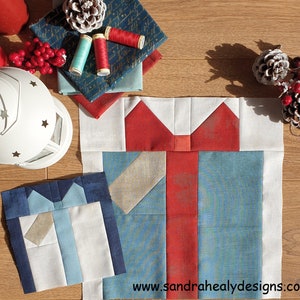 May include: A blue and white fabric gift box with a red bow. The gift box is made of patchwork fabric squares. The fabric squares are sewn together to create a pattern. The gift box is on a wooden surface.