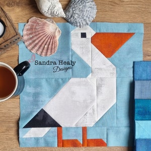May include: A fabric block featuring a geometric design of a pelican in shades of white, gray, orange, and blue. The block is set against a light blue background, with the text "Sandra Healy Designs" in cursive. A seashell and a cup of tea are also visible.