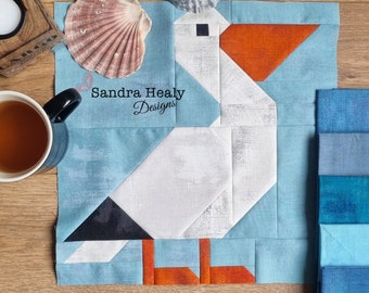 Può includere: Un blocco di tessuto patchwork con un disegno geometrico di un pellicano nei toni del bianco, grigio, arancione e blu. Il blocco è ambientato su uno sfondo azzurro, con il testo "Sandra Healy Designs". Sono visibili anche una conchiglia e una tazza di tè.