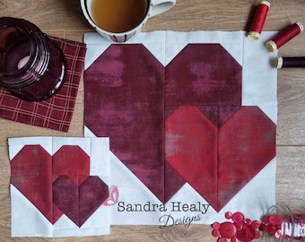 May include: Two quilt blocks featuring a heart design in shades of red and burgundy. The blocks are on a white background with the text "Sandra Healy Designs" printed below.
