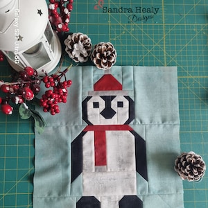 Puede incluir: Un bloque de tela turquesa con un diseño de pingüino de patchwork. El pingüino lleva un gorro rojo y una bufanda roja. El bloque de tela está sobre una estera de corte verde.