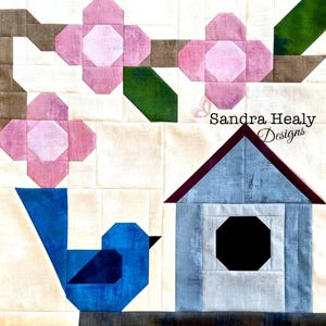 Puede incluir: Un pájaro azul con un ojo marrón se posa en una rama con flores rosas. Una casita de pájaros con un techo burdeos y una abertura circular negra está en el fondo. El texto "Sandra Healy Designs" está en la esquina superior derecha de la imagen.
