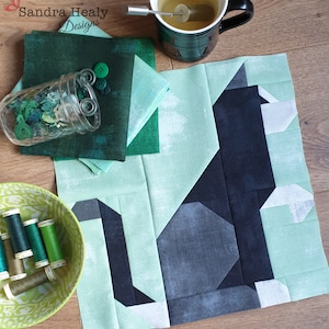 May include: A quilt block with a black cat design on a light green background. The cat is made up of different shades of green and black fabric. The quilt block is surrounded by a border of light green fabric.