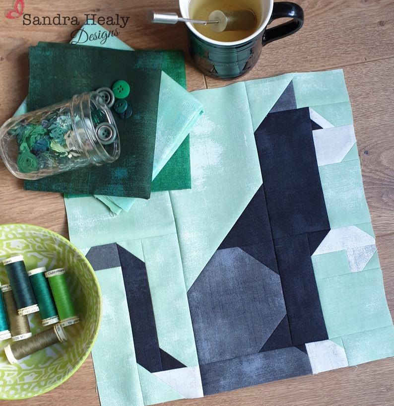 May include: A quilt block with a black cat design on a light green background. The cat is made up of different shades of green and black fabric. The quilt block is surrounded by a border of light green fabric.