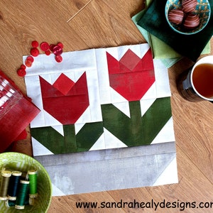 Puede incluir: Un bloque de colcha de retazos rojo y verde con dos tulipanes estilizados en una maceta gris. El bloque está hecho de cuadrados de tela cosidos juntos para crear un diseño geométrico. El fondo es blanco.