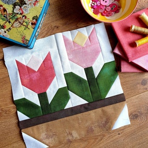 May include: A white fabric panel with two pink and red tulips in a brown pot. The tulips are made of fabric and have green leaves. The fabric panel is perfect for a quilting project.