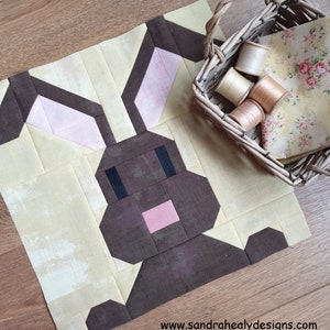 May include: A brown and pink patchwork bunny quilt block with a yellow background. The block is made of fabric squares sewn together to create the shape of a bunny. The bunny has a pink nose, brown eyes, and brown ears. The block is sitting on a wooden surface.