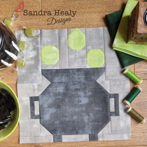 May include: A gray and green patchwork quilt block featuring a cauldron with green candy on top. The block is surrounded by green fabric scraps and spools of thread.
