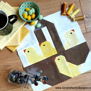 May include: A quilt block with a brown and white background featuring four yellow chicks with orange beaks and black eyes. The chicks are arranged in a nest-like shape. The quilt block is surrounded by yellow fabric scraps, a bowl of candy eggs, and spools of thread.