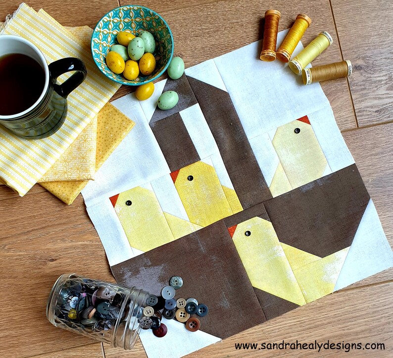 May include: A quilt block with a brown and white background featuring four yellow chicks with orange beaks and black eyes. The chicks are arranged in a nest-like shape. The quilt block is surrounded by yellow fabric scraps, a bowl of candy eggs, and spools of thread.