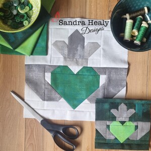 May include: A green and gray fabric quilt block with a heart shape in the center. The block is on a white background with a pair of scissors and a bowl of buttons. The text "Sandra Healy Designs" is visible in the upper right corner of the image.