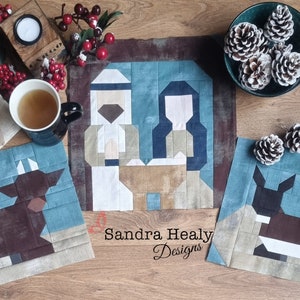 May include: Three fabric squares with a blue background and a brown, white and beige patchwork design. The centre square depicts a nativity scene with a man and woman in white and beige clothing. The left and right squares depict a brown and white animal, possibly a cow or a deer. The squares are on a wooden surface with pine cones and a cup of tea.