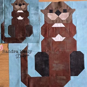Könnte beinhalten: Ein Stoff-Quiltblock-Muster, das einen braunen Otter mit einem weißen Brustfleck und dunkelbraunen Pfoten vor einem hellblauen Hintergrund zeigt. Das Design enthält den Text Sandra Healy Designs und ein kleines rosa Schmetterlingssymbol.