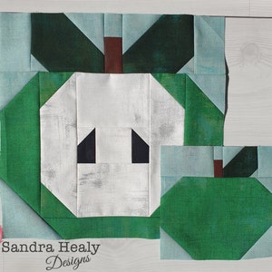 Pode incluir: Um bloco de colcha de retalhos verde e branco com um design de maçã. A maçã é feita de tecido branco com olhos pretos e uma haste marrom. A maçã é cercada por tecido verde com um fundo azul.