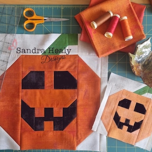 May include: Two unfinished patchwork pumpkin quilt blocks with a black and orange color scheme. The larger block has the text "Sandra Healy Designs" in white.