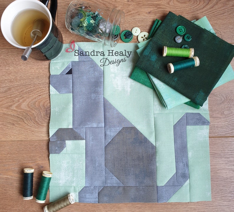 May include: A quilt block featuring a gray stylized dog design on a green background. The block is surrounded by green fabric scraps and spools of thread. The text "Sandra Healy Designs" is visible in the upper center of the image.
