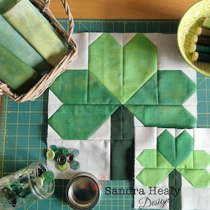 May include: A green and white fabric quilt block with a four-leaf clover design. The block is made of squares and triangles of green fabric, with white fabric in between. The block is surrounded by a white border. The text "Sandra Healy Designs" is printed in black at the bottom of the block.