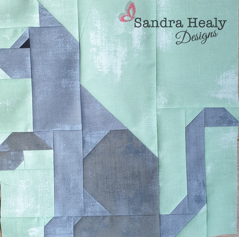May include: A quilt block featuring a gray cat design with a light green background. The cat is made up of geometric shapes and the background has a subtle texture. The text "Sandra Healy Designs" is in the upper right corner of the block.