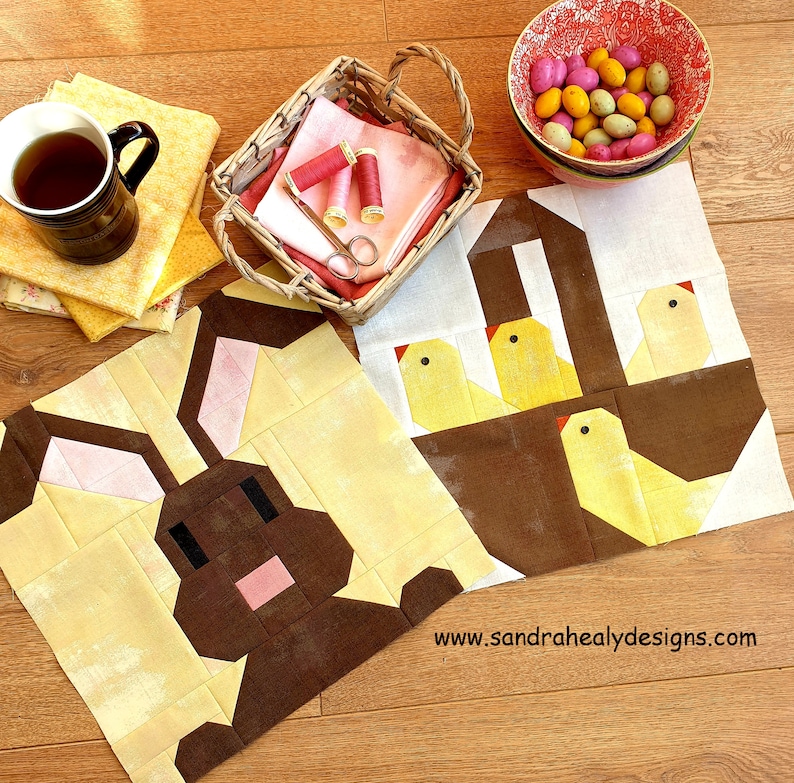 May include: Two fabric squares with a patchwork design. One square features a brown bunny with pink ears and the other square features a brown basket with three yellow chicks. The text "www.sandrahealydesigns.com" is printed on the bottom right corner of the image.