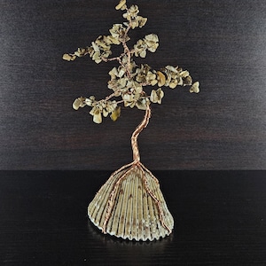 Könnte beinhalten: Eine handgefertigte Bonsai-Baum-Skulptur. Der Baum hat einen Stamm und Äste aus Kupferdraht, verziert mit kleinen, hellgrünen Steinen. Die Basis ist eine große, strukturierte Muschel, die eine einzigartige und natürliche Ästhetik schafft.