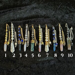 May include: A collection of ten crystal pendulums, each with a unique design. They feature various gemstones, beads, and chains, with numbers 1 through 10 displayed below. The pendulums are arranged on a dark, textured surface.
