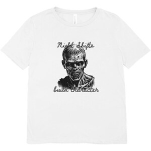 May include: A white short-sleeved t-shirt featuring a black and white graphic illustration of a zombie face in the center. Above the graphic is the text Night Shifts and below the graphic is the text build character in a cursive script font.