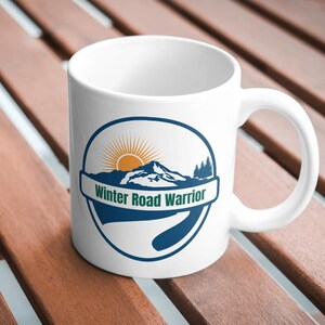 May include: A white ceramic mug featuring a circular graphic with a blue border. The design includes a stylized orange sun rising over a dark blue mountain range with pine trees and a winding road. The text Winter Road Warrior is printed in green.