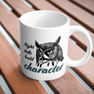 May include: A white ceramic mug featuring a black and white illustration of an owl's head. The text "Night shifts build character" is printed on the front in a combination of black serif and dark blue script fonts, centered on the mug.