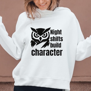 May include: A white long-sleeve hooded sweatshirt featuring a black graphic of an owl head and the text "Night shifts build character" printed on the front. The garment has a hood, long sleeves with cuffs, and a straight hemline at the waist.
