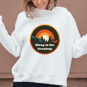 May include: A white long-sleeve hooded sweatshirt featuring a circular graphic on the chest. The design displays a sunset over a forest silhouette with the text Sleep is for breakup in a bold, dark font centered within the orange and black circle.