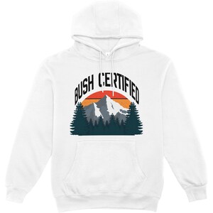 May include: A white hooded sweatshirt featuring a graphic print of a mountain range and pine trees set against a semi-circular orange and red sunset. The text BUSH CERTIFIED is printed in a bold black sans-serif font in an arch above the graphic.