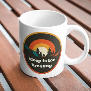 May include: A white ceramic mug featuring a circular graphic with a sunset landscape of mountains and pine trees in shades of orange, yellow, and dark blue. The text "Sleep is for breakup" is printed in white letters at the bottom of the graphic.