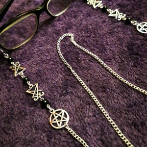 May include: A silver-toned eyeglass chain featuring alternating black beads, pentagram charms, and sigil charms. The chain is attached to black-framed eyeglasses, with a long, thin silver metal link chain extending from the decorative segments.