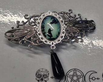 Op de afbeelding: Een zilverkleurige metalen haarspeld met een ovale cabochon met een silhouetillustratie van een meisje en een kat tegen een lichtblauwe achtergrond. De speld heeft gedetailleerd filigraanwerk en een bungelende zwarte traanvormige kraal.