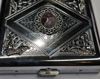 Könnte beinhalten: Ein quadratisches, silberfarbenes Metallgehäuse mit einem geprägten Rautenmuster auf dem Deckel. In der Mitte befindet sich ein ovaler, violett schimmernder Stein in einer filigranen Fassung. Jede Ecke des Deckels ist mit einer kleinen Fledermaus verziert.