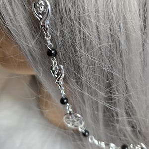 May include: A silver-toned headband featuring a series of small, black beads and decorative charms. The charms include winged skulls and a pentagram, creating a gothic or alternative aesthetic. The headband is set against a backdrop of gray hair.