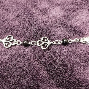 May include: A silver chain bracelet with two black beads and decorative silver charms. The charms are in the shape of a stylized heart.