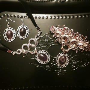 May include: A jewellery set consisting of a pair of silver-toned earrings, a choker necklace, and a hair clip. Each piece features a silver-toned metal frame with intricate filigree detailing and a central oval-shaped dark purple cabochon stone.