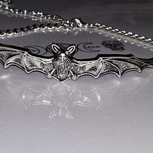 Può includere: Una collana in metallo color argento con un ampio e dettagliato ciondolo a forma di pipistrello con ali spiegate. Il pipistrello presenta dettagli di pelliccia testurizzati e orecchie a punta. Il ciondolo è fissato a una semplice catena.