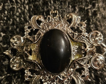 May include: A silver-toned metal brooch featuring an oval-shaped black cabochon stone at the center. The frame is decorated with intricate filigree scrollwork and symmetrical patterns, creating a detailed ornamental design around the central stone.