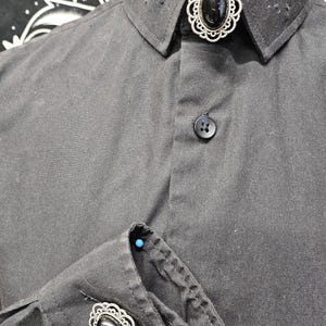 May include: A black button-down shirt with a silver collar pin featuring a black oval stone. The shirt has a silver cufflink with a black oval stone on the left cuff.