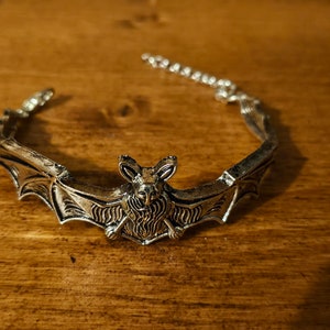 May include: A silver bat-shaped bracelet with a detailed design. The bracelet is open and has a chain link closure.