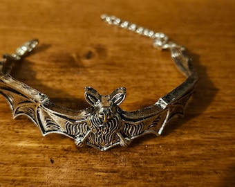 May include: A silver-toned metal bracelet featuring a central bat motif with detailed wings and fur texture. The bracelet includes a delicate chain link extension on one side, allowing for adjustable sizing, and has a weathered, antiqued finish.