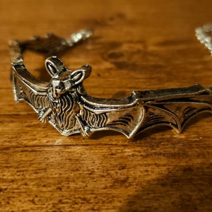 May include: Silver-toned bat-shaped bracelet with intricate details. The bat is depicted with outstretched wings and a detailed face.