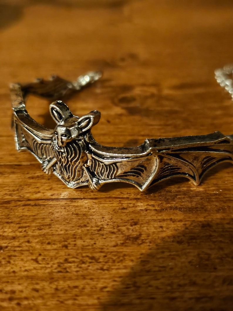 May include: Silver-toned bat-shaped bracelet with intricate details. The bat is depicted with outstretched wings and a detailed face.