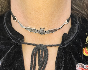 Może przedstawiać: Wisiorek w kształcie nietoperza wykonany ze srebrzystego metalu, umieszczony na cienkim czarnym sznurku typu choker. Nietoperz jest przedstawiony z szeroko rozpostartymi skrzydłami, z wyraźną fakturą skrzydeł i małą głową.