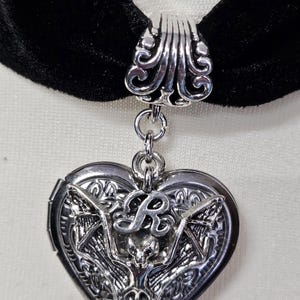 Peut inclure: Un collier ras du cou en velours noir avec un pendentif argenté en forme de cœur. Le pendentif présente un motif d'ailes de chauve-souris, un crâne et la lettre "R". Le collier est un accessoire de style gothique.