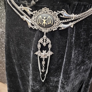 May include: A silver-toned chain belt featuring a central oval medallion with a gold bat silhouette on a black background. The belt includes silver hand-shaped connectors, decorative filigree metalwork, a hanging bat charm, and a black crystal pendant.
