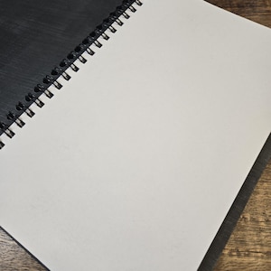 May include: An open spiral-bound notebook with a black cover and blank white pages. The notebook is lying on a wooden surface, with the pages open to reveal the clean, unlined paper. The spiral binding is visible along the left edge.